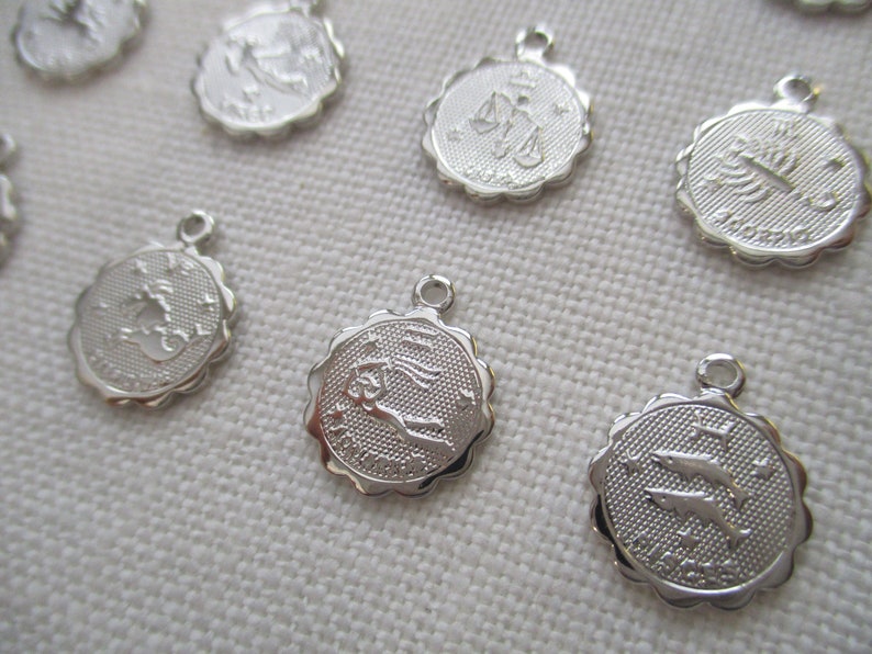 May include: A collection of silver-colored zodiac charms. Each charm features a different zodiac symbol, such as scales, a scorpion, and a pair of fish, with a textured background and a scalloped edge. Each charm has a small loop at the top.