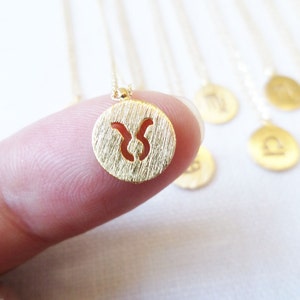 May include: A gold-colored necklace featuring a small, round pendant with a textured surface and a cut-out Taurus zodiac symbol. The necklace is held by a finger, with other zodiac pendants visible in the background.