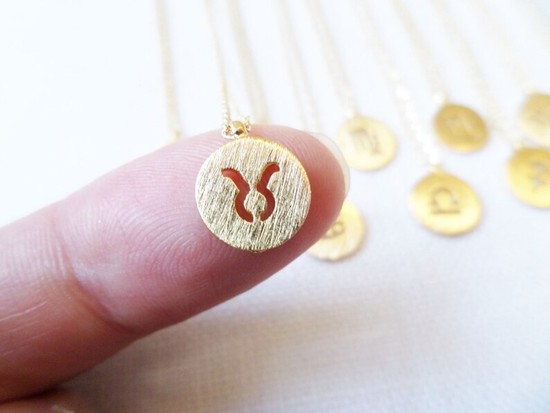 May include: A gold-colored necklace featuring a small, round pendant with a textured surface and a cut-out Taurus zodiac symbol. The necklace is held by a finger, with other zodiac pendants visible in the background.
