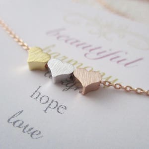 May include: A delicate gold, silver, and rose gold necklace with three small heart charms. The necklace is on a white card with the words "beautiful harmony hope love" written on it.