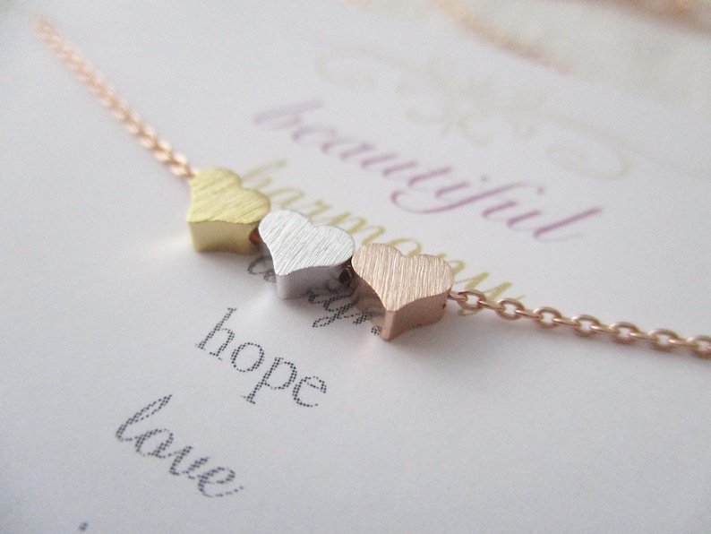 May include: A delicate gold, silver, and rose gold necklace with three small heart charms. The necklace is on a white card with the words "beautiful harmony hope love" written on it.