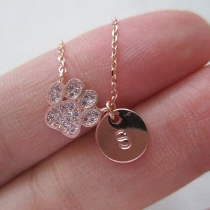 May include: Rose gold necklace featuring a paw print pendant encrusted with small, clear crystals. A round charm with the letter "S" is also attached. The delicate chain and pendants are rose gold in color, creating a simple yet elegant design.