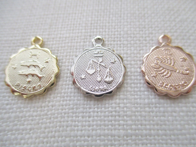 May include: Three small, round zodiac charms. One is gold with the Pisces symbol, another is silver with the Libra symbol, and the third is rose gold with the Scorpio symbol. Each charm has a scalloped edge and a small loop for attaching to a chain.