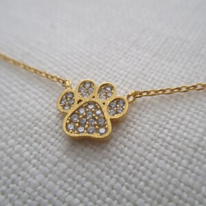 May include: A gold-colored necklace featuring a paw print pendant. The paw print is encrusted with small, clear gemstones. The necklace chain is delicate and also gold-colored. The background is a light, textured fabric.