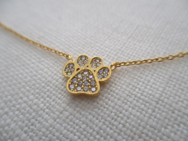May include: A gold-colored necklace featuring a paw print pendant. The paw print is encrusted with small, clear gemstones. The necklace chain is delicate and also gold-colored. The background is a light, textured fabric.