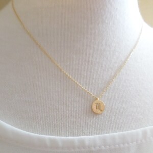 May include: A delicate gold-colored necklace featuring a small, round pendant. The pendant displays a stylized symbol, likely a zodiac sign. The necklace is shown against a white background, highlighting the jewelry's simple elegance.