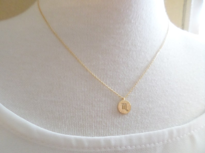 May include: A delicate gold-colored necklace featuring a small, round pendant. The pendant displays a stylized symbol, likely a zodiac sign. The necklace is shown against a white background, highlighting the jewelry's simple elegance.