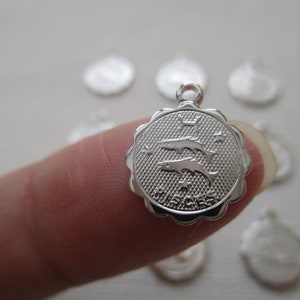 May include: A silver-colored Pisces charm with a scalloped edge. The charm features two dolphins and the word "PISCES" engraved on a textured background. The charm has a small loop at the top for attaching to a chain or bracelet.
