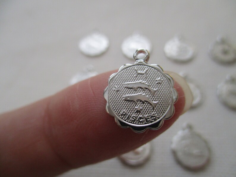 May include: A silver-colored Pisces charm with a scalloped edge. The charm features two dolphins and the word "PISCES" engraved on a textured background. The charm has a small loop at the top for attaching to a chain or bracelet.