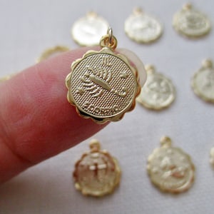 May include: A gold-colored pendant with a scalloped edge, featuring the Scorpio zodiac symbol and the word "SCORPIO". The pendant is held by a finger, with other similar pendants in the background. The pendant is small and round.