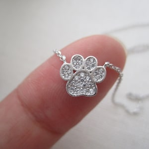 May include: A silver-colored necklace featuring a paw print pendant. The pendant is encrusted with small, clear gemstones. The necklace chain is delicate and thin. The paw print design is a popular symbol for pet lovers.