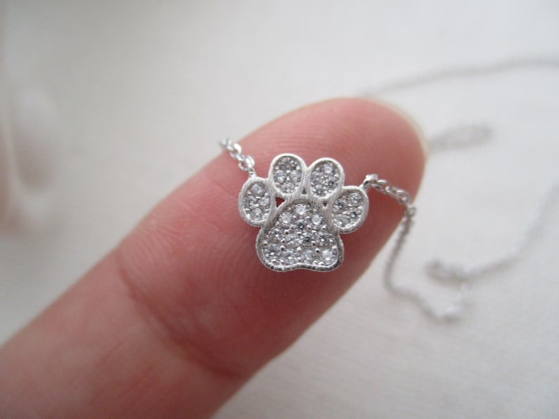 May include: A silver-colored necklace featuring a paw print pendant. The pendant is encrusted with small, clear gemstones. The necklace chain is delicate and thin. The paw print design is a popular symbol for pet lovers.