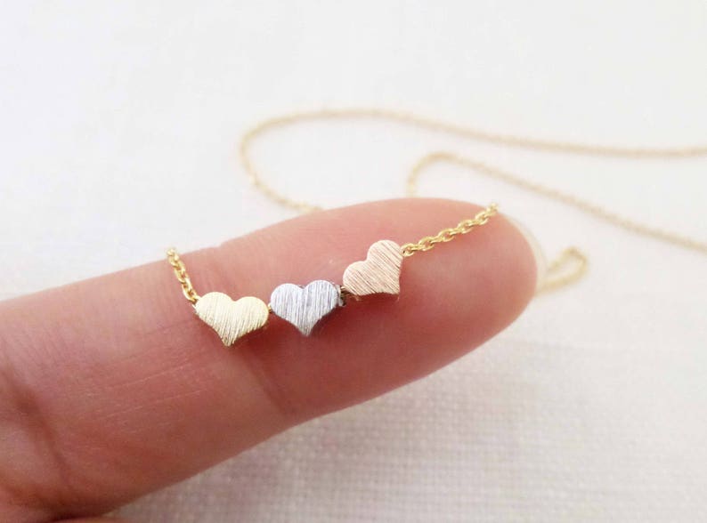 May include: A gold chain necklace with three small heart charms. The hearts are different colors: gold, silver, and rose gold.