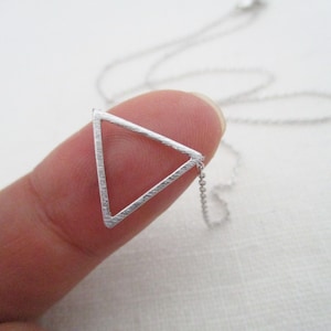 Puede incluir: Un collar con un colgante de triángulo plateado y una cadena delicada. El triángulo es abierto y tiene un acabado texturizado.