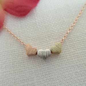 May include: A delicate gold chain necklace with three small heart charms. The hearts are layered and each has a different finish: rose gold, silver, and yellow gold.