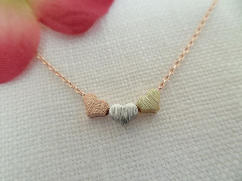May include: A delicate gold chain necklace with three small heart charms. The hearts are layered and each has a different finish: rose gold, silver, and yellow gold.