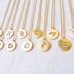 May include: A collection of gold-colored necklaces, each featuring a circular pendant with a different zodiac symbol. The pendants are suspended from delicate chains. The necklaces are displayed on a white surface.