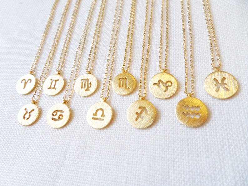May include: A collection of gold-colored necklaces, each featuring a circular pendant with a different zodiac symbol. The pendants are suspended from delicate chains. The necklaces are displayed on a white surface.