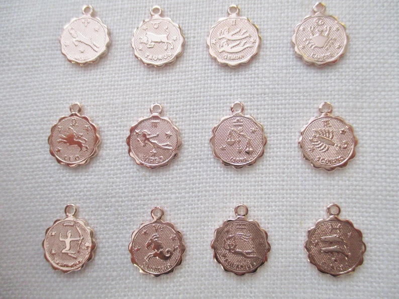 May include: Rose gold-colored zodiac charms. Each charm is round with a scalloped edge and features a different zodiac symbol and name. The charms have a small loop at the top for attaching to a chain or bracelet. The charms are displayed on a white surface.