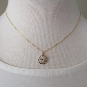 May include: A gold-colored necklace with a small, round pendant. The pendant has a scalloped edge and a design in the center. The necklace is displayed on a white background.