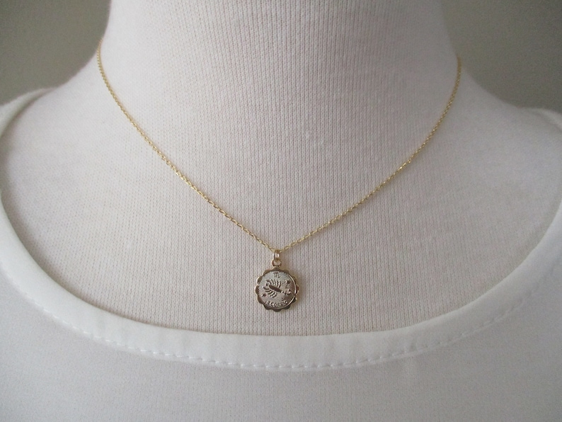 May include: A gold-colored necklace with a small, round pendant. The pendant has a scalloped edge and a design in the center. The necklace is displayed on a white background.