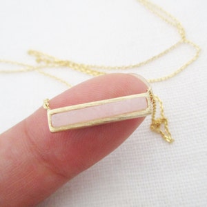 May include: A delicate gold chain necklace with a rectangular pendant featuring a light pink stone.