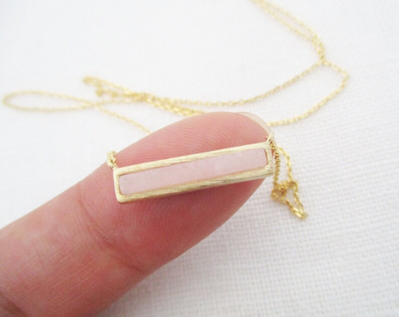 May include: A delicate gold chain necklace with a rectangular pendant featuring a light pink stone.