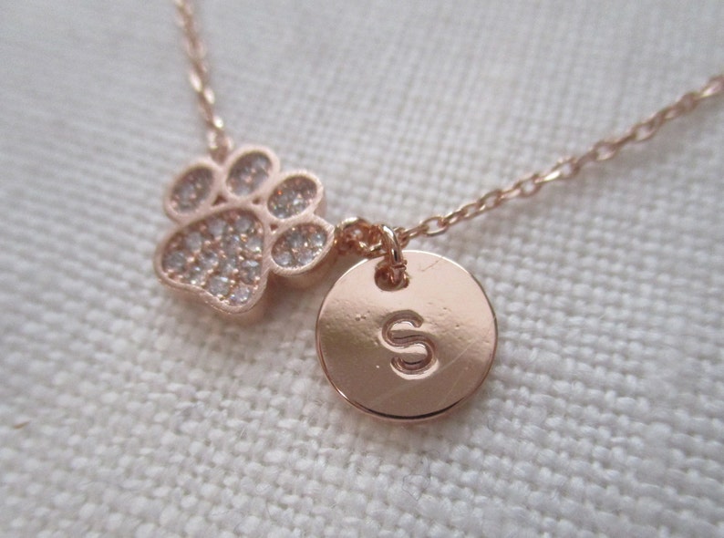 May include: Rose gold necklace featuring a paw print charm with clear crystals and a round pendant engraved with the letter "S". The delicate chain and charms are rose gold in color.