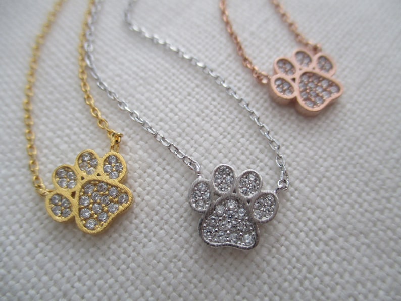 May include: Three delicate necklaces with paw print pendants. One is gold-toned, one is silver-toned, and the third is rose gold-toned. Each pendant is encrusted with small, clear crystals. The necklaces are displayed on a light-colored, textured surface.