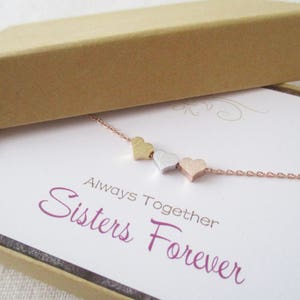 May include: A delicate gold, silver, and rose gold necklace with three small heart charms. The necklace is displayed in a box with a white card that reads "Always Together Sisters Forever".