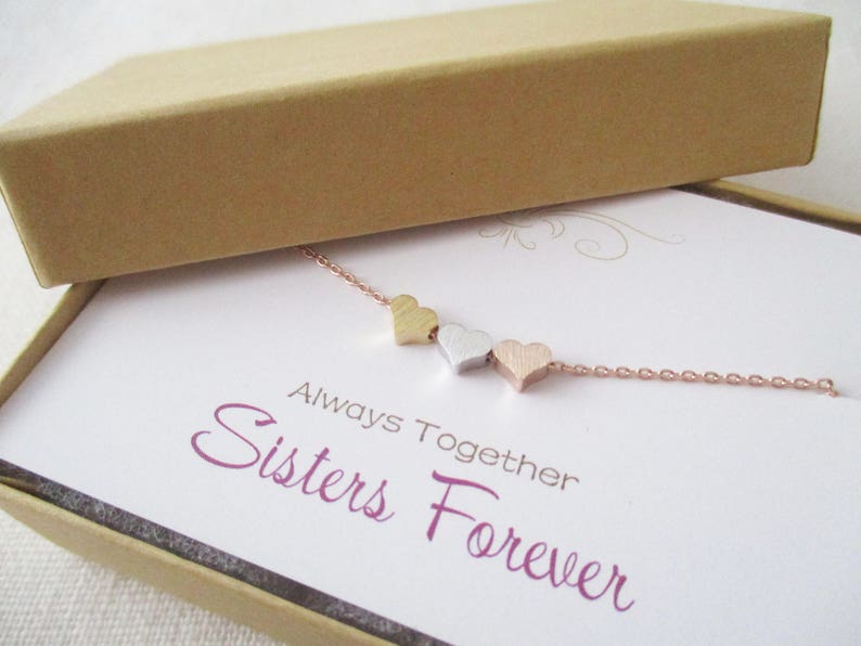 May include: A delicate gold, silver, and rose gold necklace with three small heart charms. The necklace is displayed in a box with a white card that reads "Always Together Sisters Forever".