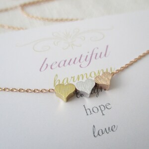 May include: A gold, silver, and rose gold heart necklace with a delicate chain. The necklace is on a white card with the words "beautiful harmony hope love" written on it.