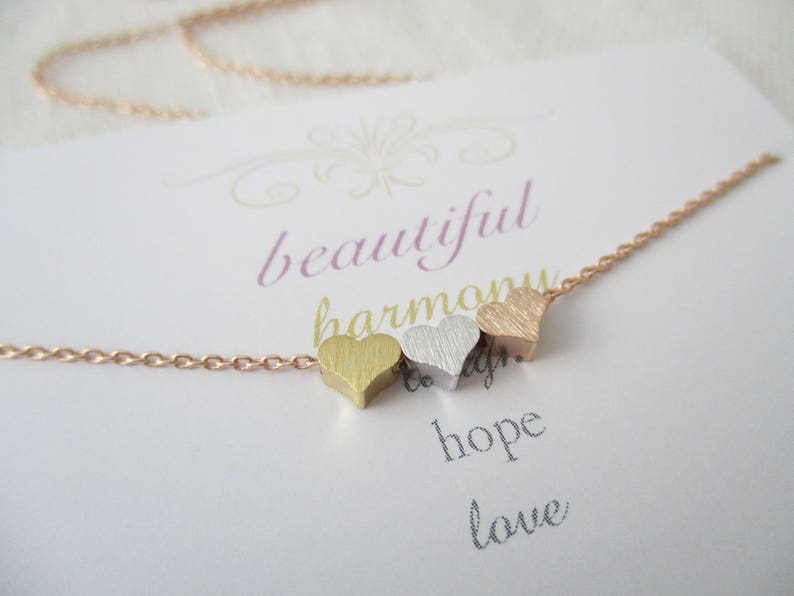 May include: A gold, silver, and rose gold heart necklace with a delicate chain. The necklace is on a white card with the words "beautiful harmony hope love" written on it.
