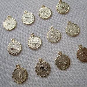 May include: A collection of gold-colored zodiac charm pendants. Each pendant features a different zodiac symbol within a scalloped edge. The charms have a small loop at the top for attaching to a chain or bracelet.