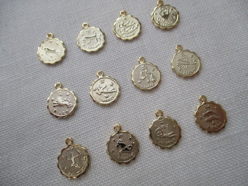 May include: A collection of gold-colored zodiac charm pendants. Each pendant features a different zodiac symbol within a scalloped edge. The charms have a small loop at the top for attaching to a chain or bracelet.