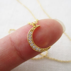 May include: A gold crescent moon necklace with clear cubic zirconia stones. The necklace is on a delicate gold chain.