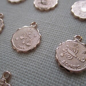 May include: Rose gold-colored zodiac charms with scalloped edges. The charms feature embossed zodiac symbols, including Libra and Scorpio, with a small loop at the top for attaching to jewelry. The background is a light fabric.