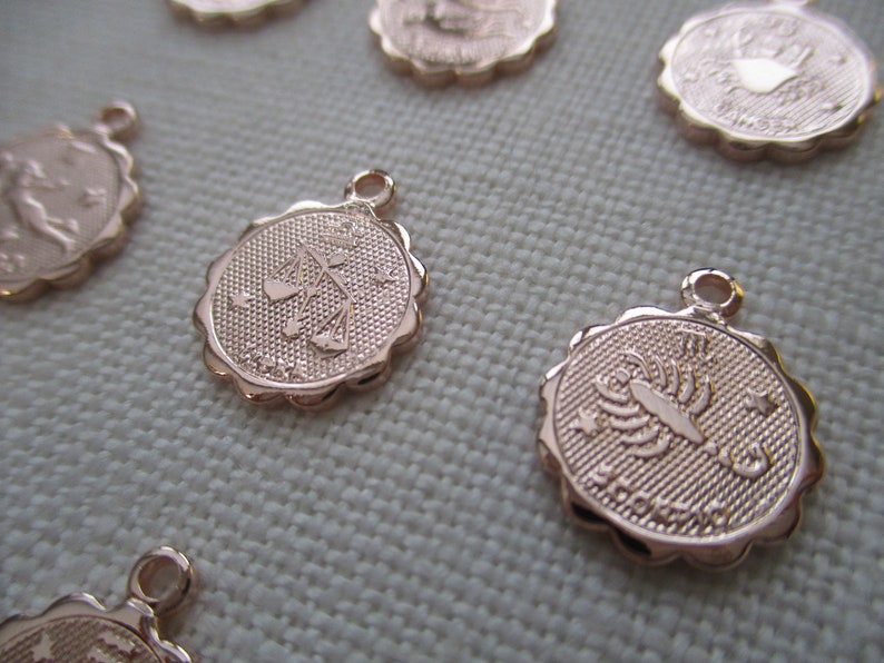 May include: Rose gold-colored zodiac charms with scalloped edges. The charms feature embossed zodiac symbols, including Libra and Scorpio, with a small loop at the top for attaching to jewelry. The background is a light fabric.