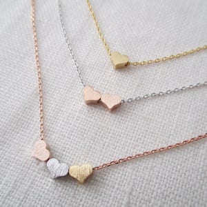May include: Three delicate gold, silver, and rose gold necklaces with heart-shaped charms. The necklaces are layered on a white fabric background.