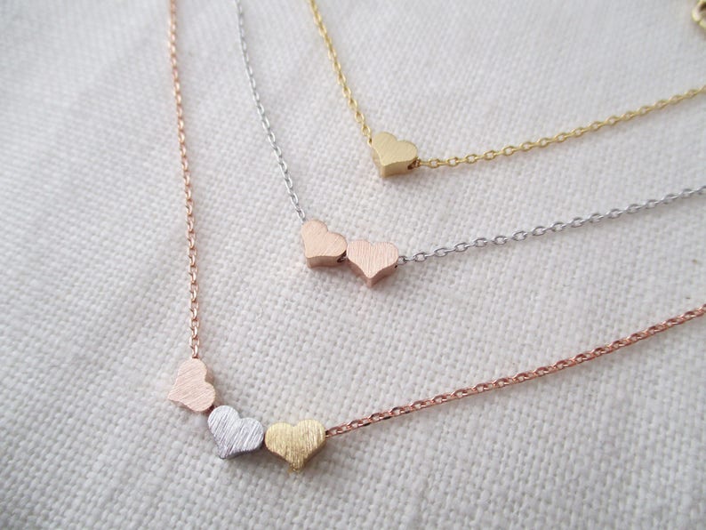 May include: Three delicate gold, silver, and rose gold necklaces with heart-shaped charms. The necklaces are layered on a white fabric background.