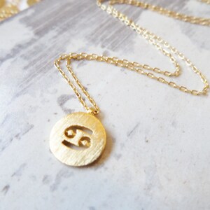 May include: Gold-colored necklace featuring a round pendant with the Cancer zodiac symbol. The pendant has a textured surface and is suspended from a delicate chain. Additional gold pendants are visible in the background.