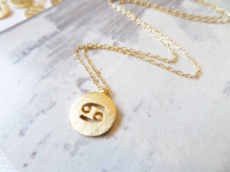 May include: Gold-colored necklace featuring a round pendant with the Cancer zodiac symbol. The pendant has a textured surface and is suspended from a delicate chain. Additional gold pendants are visible in the background.