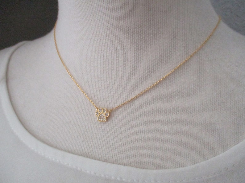 May include: A delicate gold-colored necklace featuring a small paw print pendant. The paw print is encrusted with sparkling clear crystals. The necklace is displayed on a white background.
