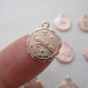 May include: A rose gold Aries zodiac charm with a scalloped edge. The charm features a ram symbol, stars, and the word "ARIES". The charm has a small loop at the top for attaching to a necklace or bracelet.