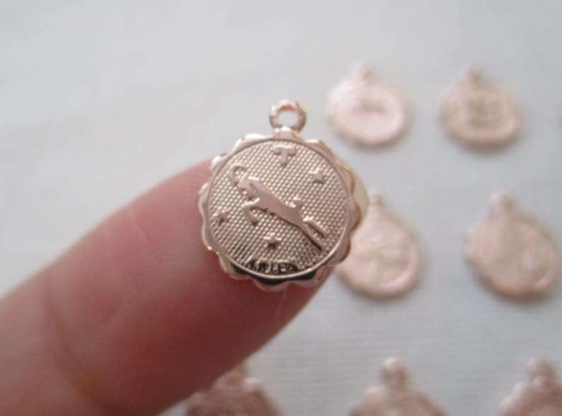 May include: A rose gold Aries zodiac charm with a scalloped edge. The charm features a ram symbol, stars, and the word "ARIES". The charm has a small loop at the top for attaching to a necklace or bracelet.
