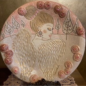Puede incluir: Plato de cerámica decorativo con una mujer con alas de ángel, rodeada de rosas rosas y árboles. El plato tiene un fondo rosa claro y blanco con detalles dorados. La mujer tiene un pendiente de flor y un accesorio para el pelo de perlas.