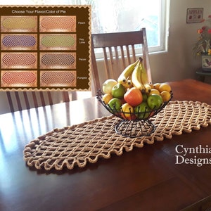 May include: A brown crocheted table runner with a diamond pattern and scalloped edges sits on a wooden table with a bowl of fruit in the center. The bowl contains bananas, apples, limes, and oranges. The text "Cynthia's Designs" is visible in the lower right corner of the image.