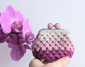 May include: A small crochet coin purse featuring a gradient of pink and purple yarn in a cluster stitch pattern. The purse has a metallic bronze-toned frame with two round, light pink clasp beads at the top, showcasing a textured handmade design.