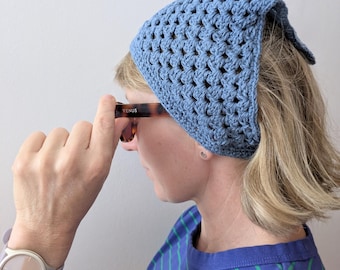 May include: A dusty blue crochet headscarf featuring a triangular shape and a textured, open-work stitch pattern. The headscarf is constructed from yarn and covers the top of the head, extending towards the back with a pointed edge.