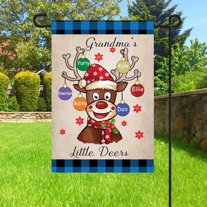 May include: A Christmas-themed garden flag with a cartoon reindeer wearing a Santa hat and a scarf. The reindeer has ornaments with names on them, including "Grandma's Little Deers", "Ellie", "Dax", "Bubby", "Emerson", "Yuri", and "Kyle".